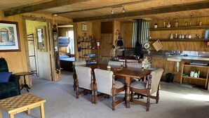 A cozy cabin interior with a wooden dining table, four chairs, a small wooden table, a blue armchair, and a kitchen area with shelves and a stove. - Riverside Retreat   (Waimakariri District)