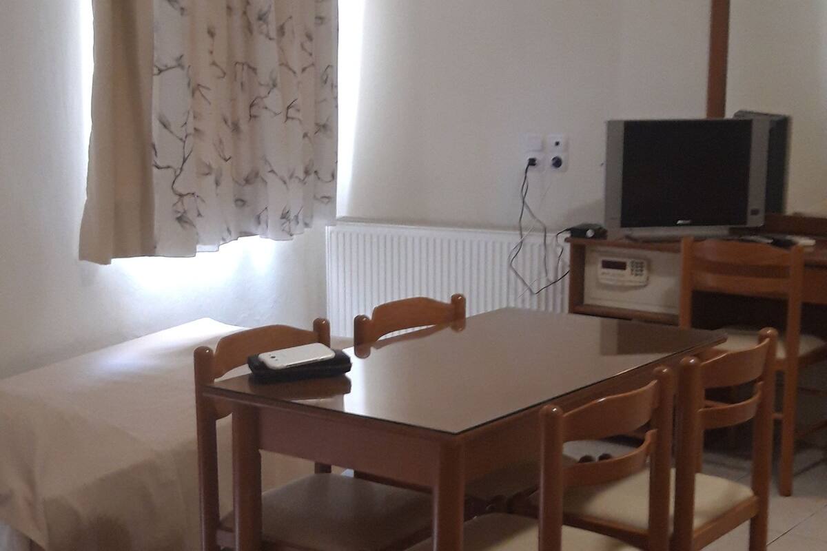 A room with a wooden dining table, four chairs, a bed with a white cover, a television on a wooden stand, and a window with curtains.