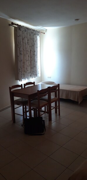 A room with a dining table and chairs, a bed with a white sheet, and a window with sheer curtains. - Dabasis-Drolias (Skiathos)