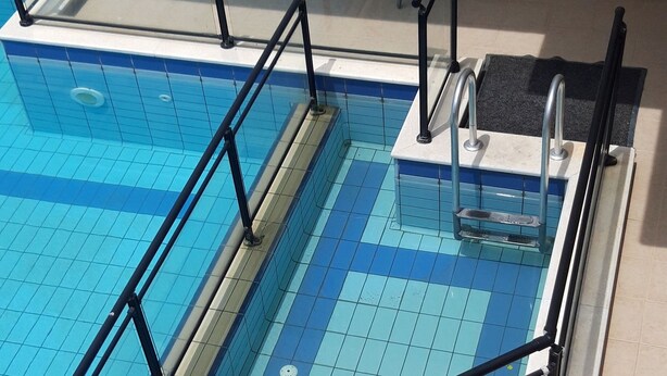 A swimming pool with a glass barrier and metal railings, a door with a number, and a small table with chairs. - Dabasis-Drolias (Skiathos)