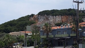 A parking lot with several cars, a building labeled "LA PISCINE," and a hillside with greenery in the background. - Dabasis-Drolias (Skiathos)