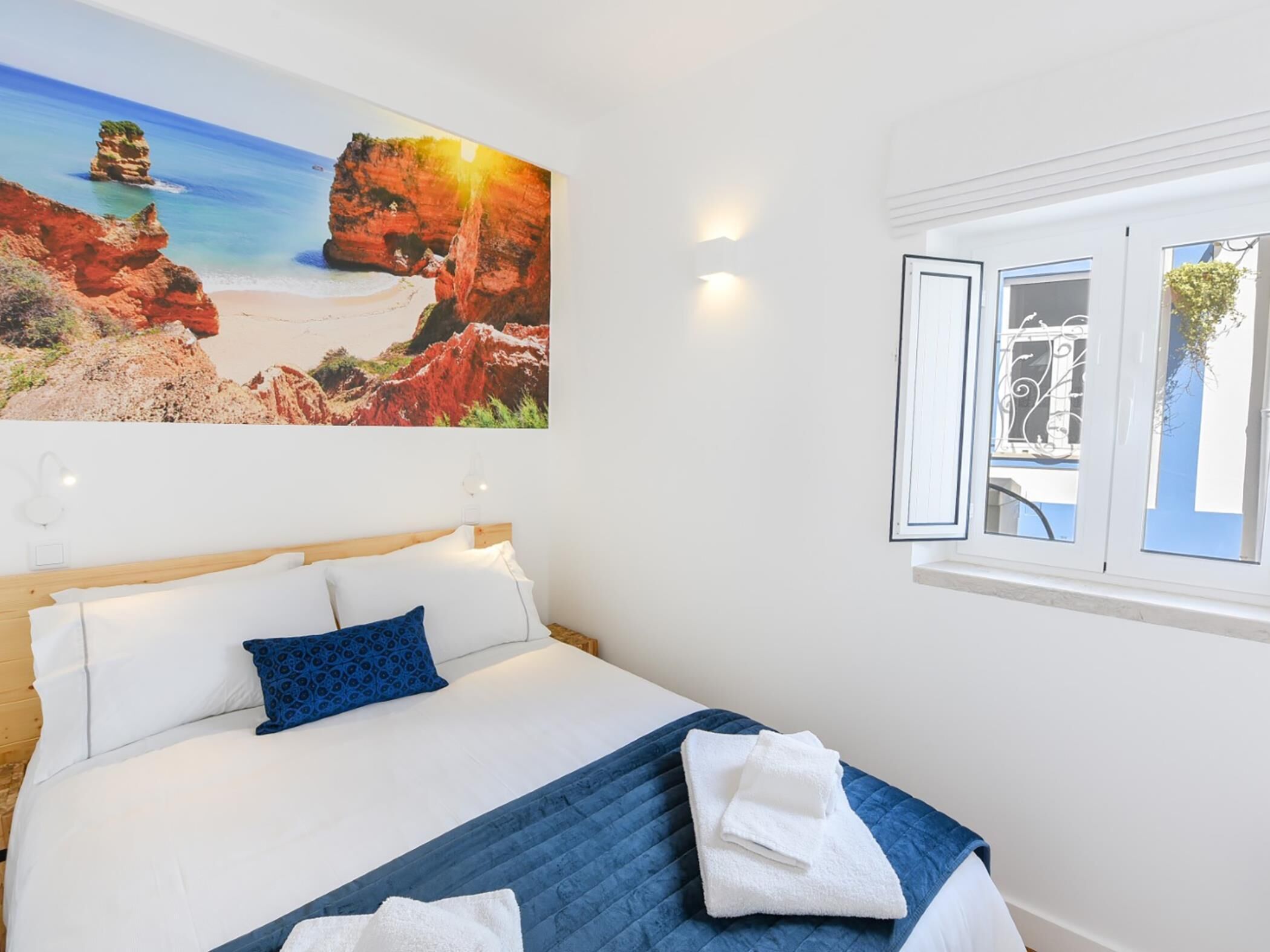 A bedroom with a bed, pillows, a quilted blue blanket, and a painting of a coastal landscape.