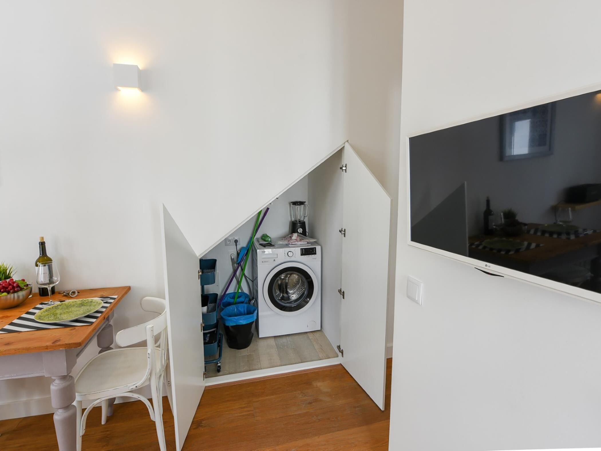 A compact kitchen with a washing machine, a dining table with chairs, and a TV mounted on the wall.