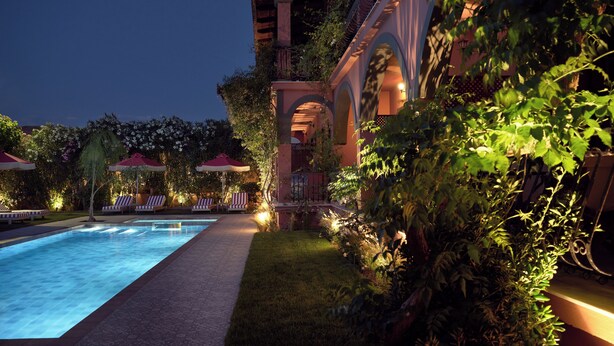 A poolside area with lounge chairs and umbrellas, surrounded by lush greenery and a building with arches. - Anemelia Retreat (Zakynthos)