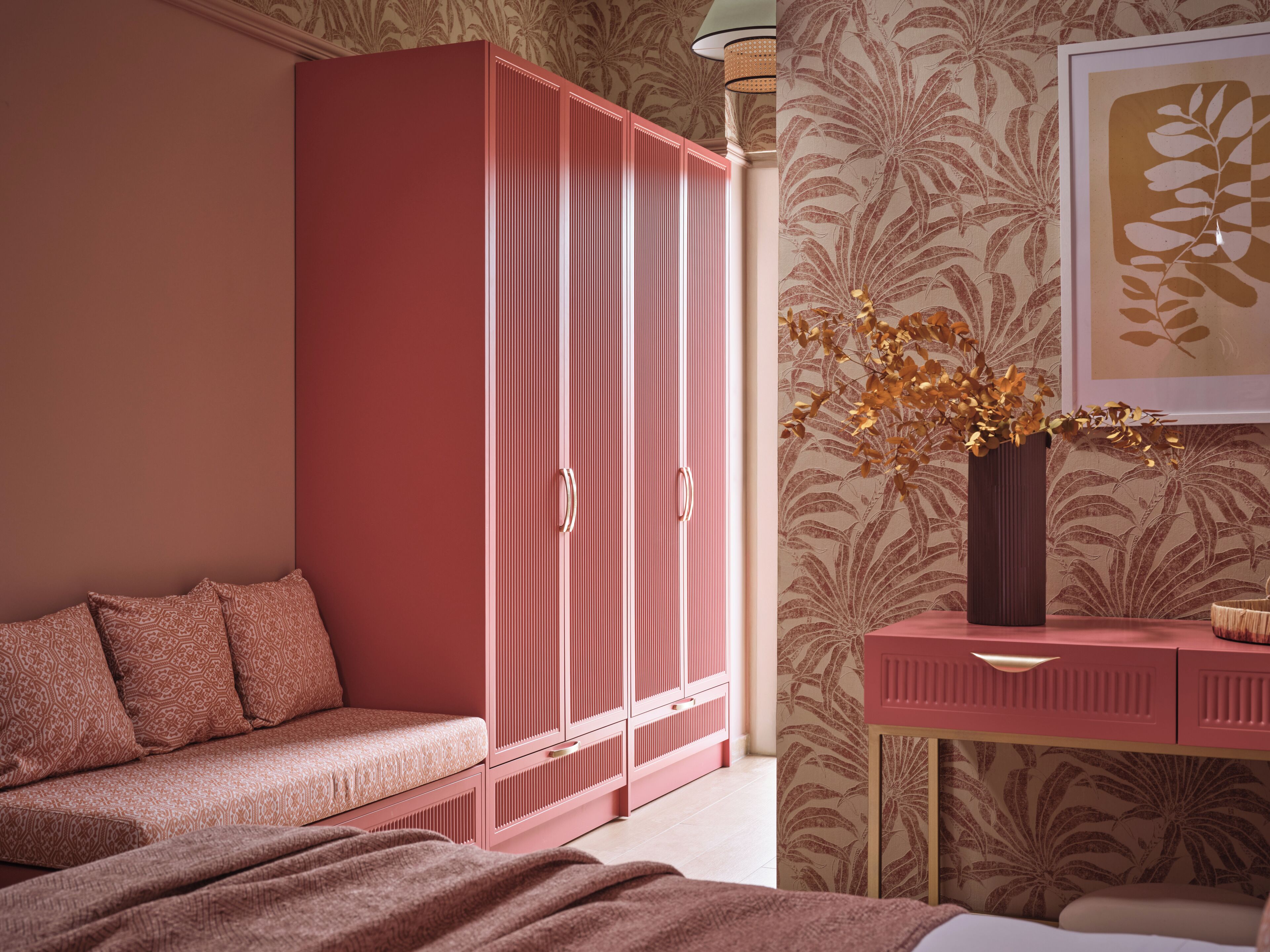 Un dormitorio con un armario rosa, una mesita de noche con un florero, papel tapiz estampado y un cuadro en la pared.