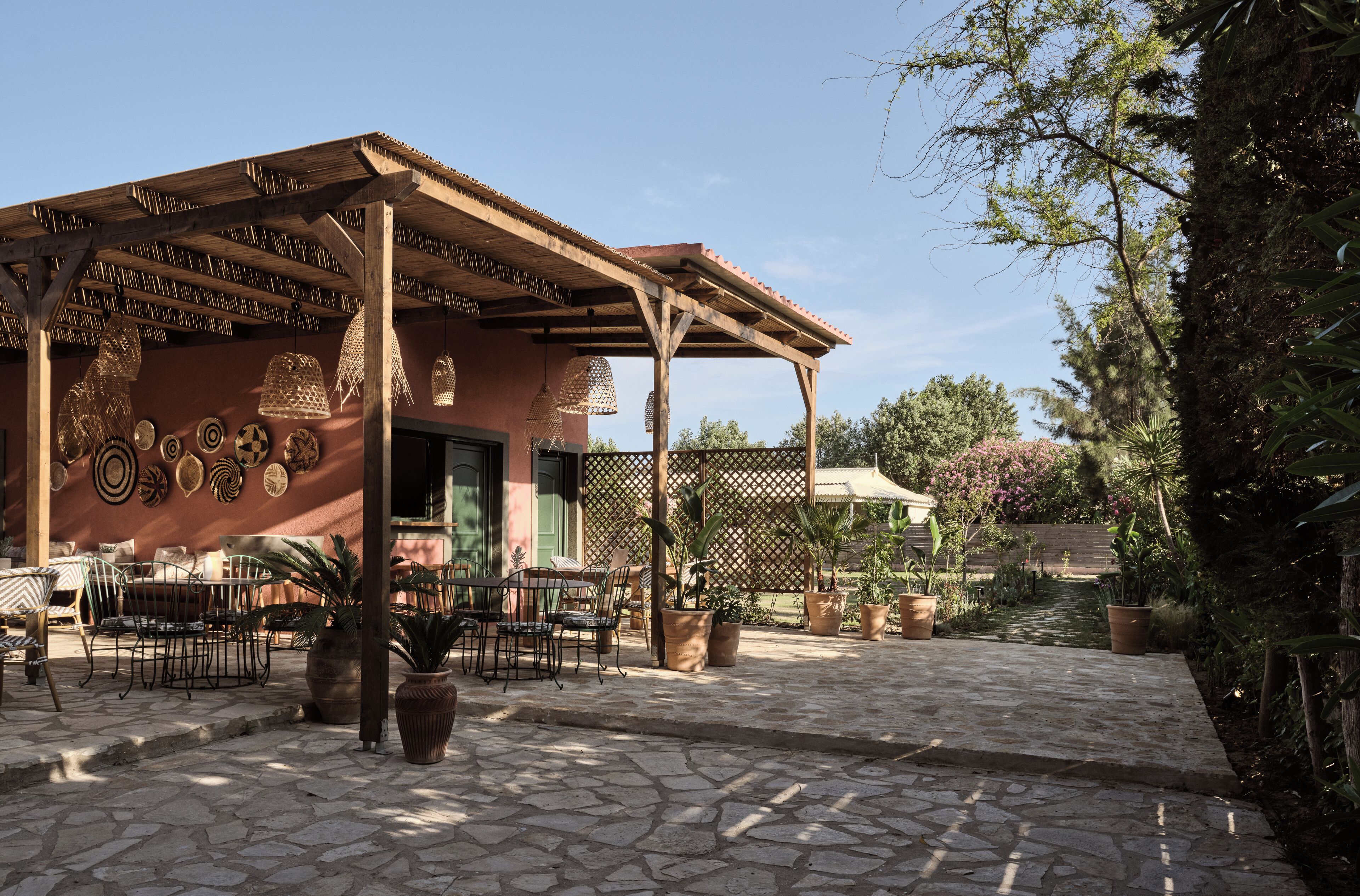 Un patio al aire libre con suelo de piedra, sillas de metal, plantas en macetas y una pérgola de madera.