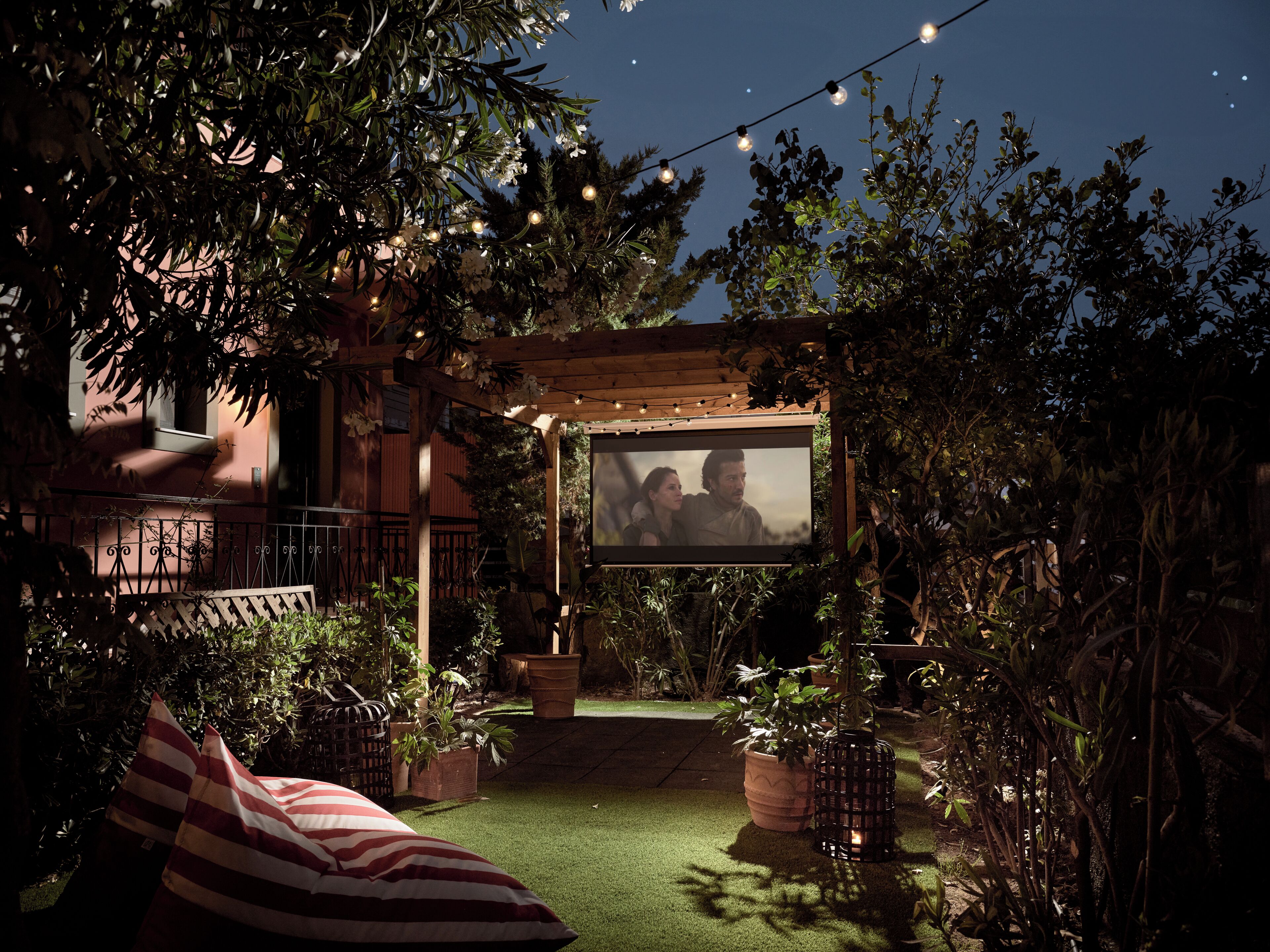 Et udendørs lærred til film i en have med lyskæder, omgivet af plantekrukker og en træpergola.
