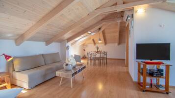 Un'ampia zona giorno con soffitto in legno a vista, un divano, un tavolo da pranzo con sedie, un televisore su un mobile e un tavolino con oggetti decorativi.