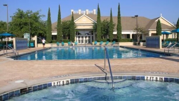 Une piscine dont la profondeur est indiquée à 2 pieds et 9 pouces, entourée de chaises longues, avec un bâtiment au fond présentant un style architectural classique.