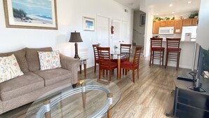Un salon avec un canapé marron, une table basse en verre et un coin repas comprenant des chaises en bois et une table en verre. Il y a également une cuisine avec des placards en bois et un four à micro-ondes.