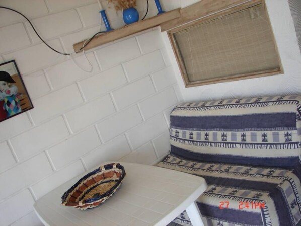 A corner of a room with a white brick wall, a small table with a woven basket, a patterned cushion, and a picture frame.