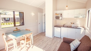 A modern kitchen with white cabinets, a microwave, and a sink. There's a dining area with a blue table and wooden chairs. A brown sofa with a striped pillow is in the living area. - Matakana Oasis - Beauregard Cottages (Matakana)