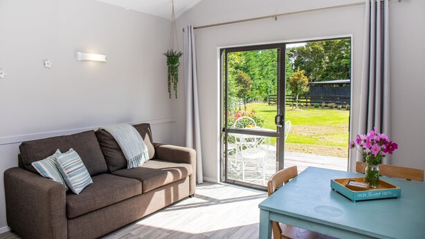 A modern living room with a brown sofa, a table with a vase of flowers, and a view of a garden through a sliding glass door. - Matakana Oasis - Beauregard Cottages (Matakana)