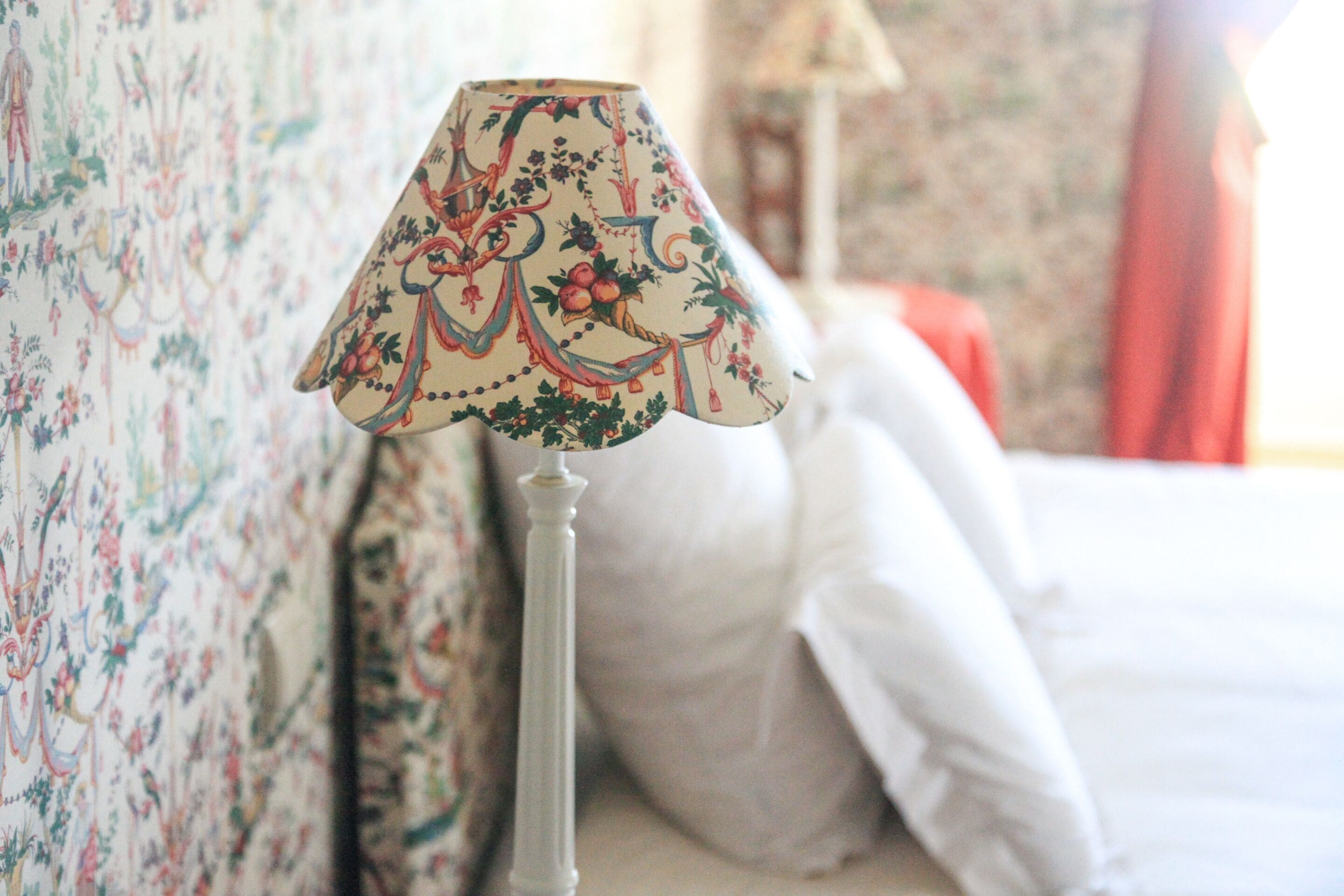 Una lámpara con estampado floral sobre un soporte blanco, ubicada junto a una cama con ropa blanca y un papel tapiz con diseño floral.