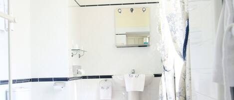 Un baño con piso de madera, sanitarios blancos, una alfombra de baño azul y una cortina de ducha estampada con motivos florales.