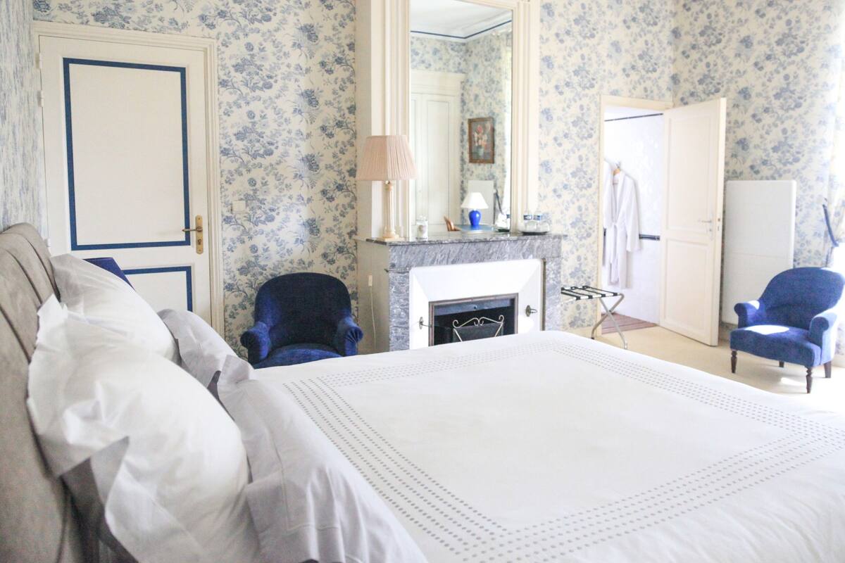 A bedroom with a large bed, a blue armchair, a chair by the fireplace, and a wardrobe.