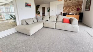 A modern living room with a sectional sofa, a small kitchen area, and a dining table. - Waitarere retreat - on the dunes, panoramic sea views, minutes to the sand (Waitarere Beach)