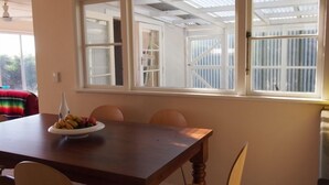 A dining area with a wooden table, chairs, and a bowl of fruit. There is a window with a view of a building and a door. - Waitarere retreat - on the dunes, panoramic sea views, minutes to the sand (Waitarere Beach)