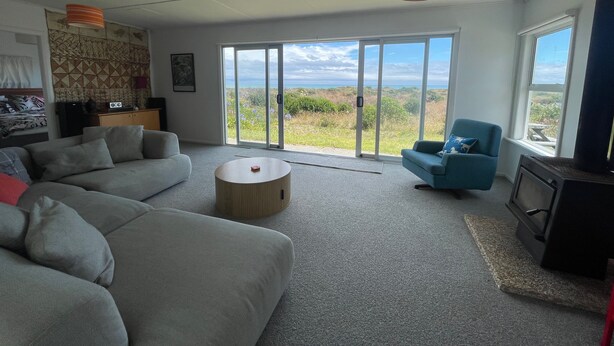 A modern living room with a grey sofa, a blue armchair, a round coffee table, and a television on a stand. - Waitarere retreat - on the dunes, panoramic sea views, minutes to the sand (Waitarere Beach)