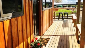 Un'area esterna in legno con panchina e tavolo, una pianta in vaso con fiori rossi, e sullo sfondo un prato verde e alberi.