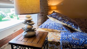 A bed with blue floral bedding, a lamp with a woven shade, and a stack of stones on a wooden table. - Aux Mésanges (Labergement-Foigney)