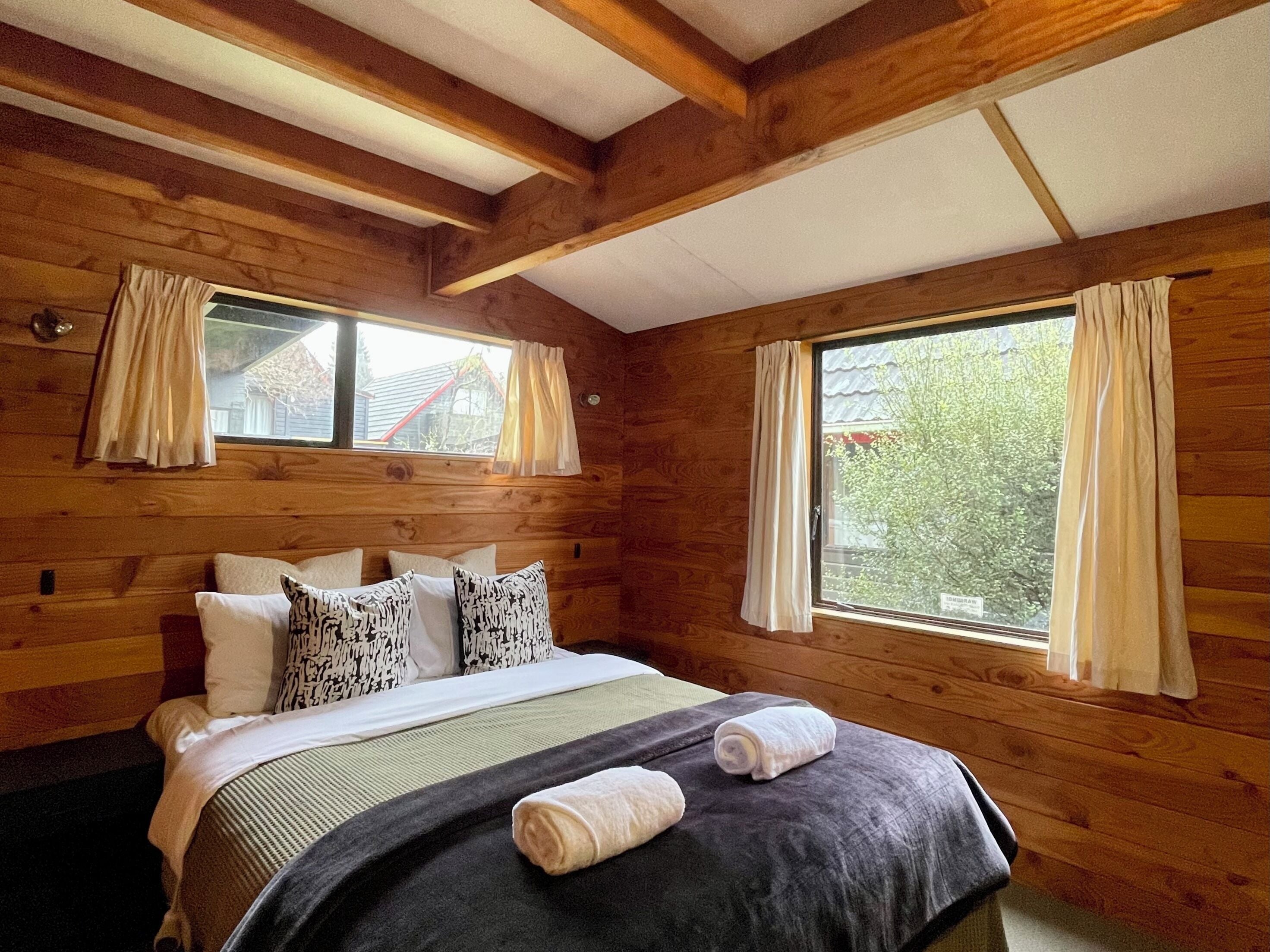 A wooden cabin room with a bed, two windows, and curtains.