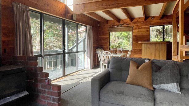 A cozy cabin interior with a fireplace, a gray sofa with cushions, a dining area with chairs, and large windows offering views of the outdoors. - Snow Chalet - Ohakune Chalet (Ohakune)