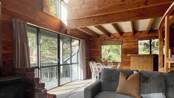 A cozy cabin interior with a fireplace, a gray sofa with cushions, a dining area with chairs, and large windows offering views of the outdoors.