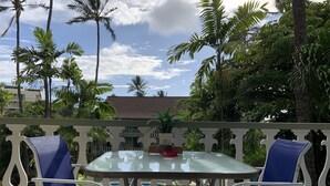 A glass-top table with two blue cushioned chairs on a balcony with white railings, surrounded by lush greenery and a clear sky. - RENOVATED Kona Studio By The Ocean!!! (Kailua)