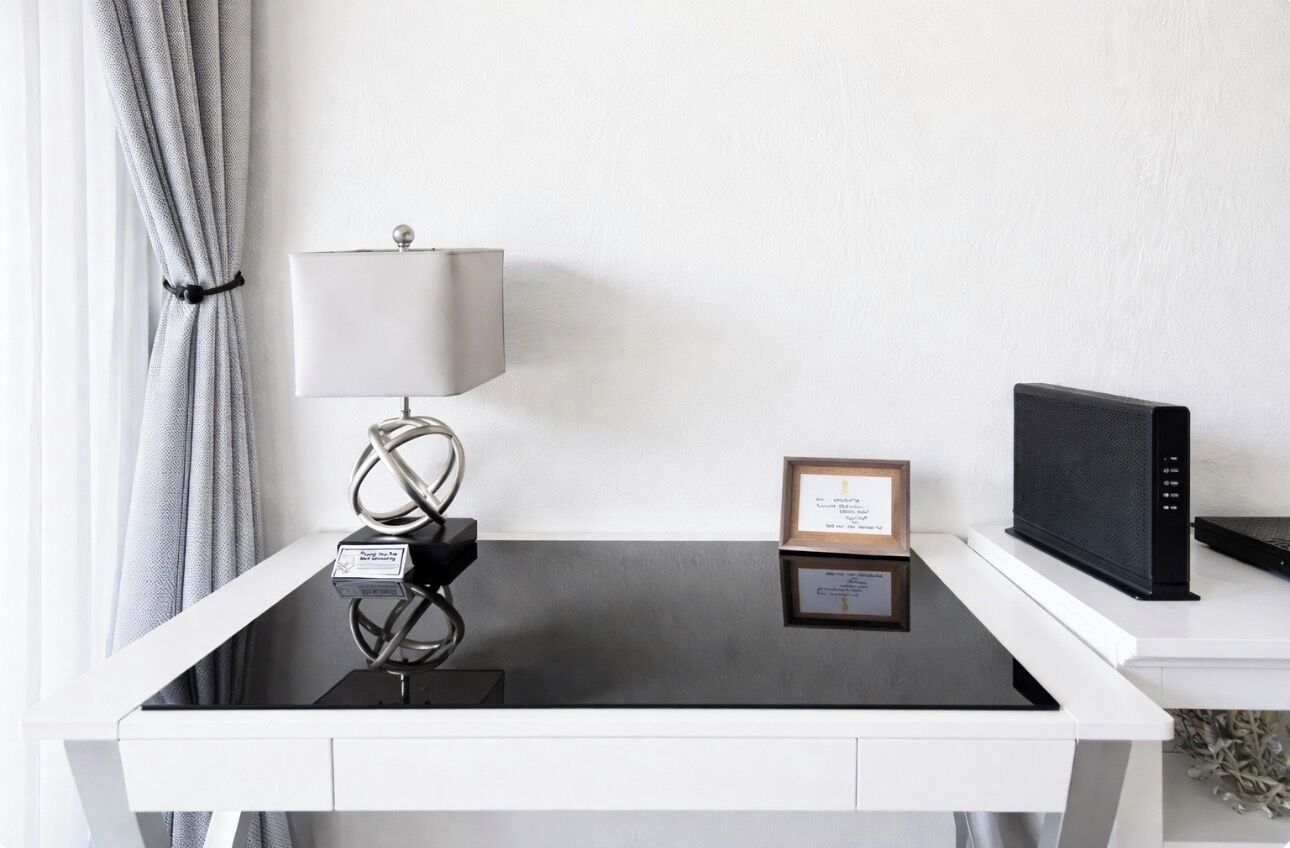 A modern desk with a lamp, a framed certificate, and a black speaker on a white table.