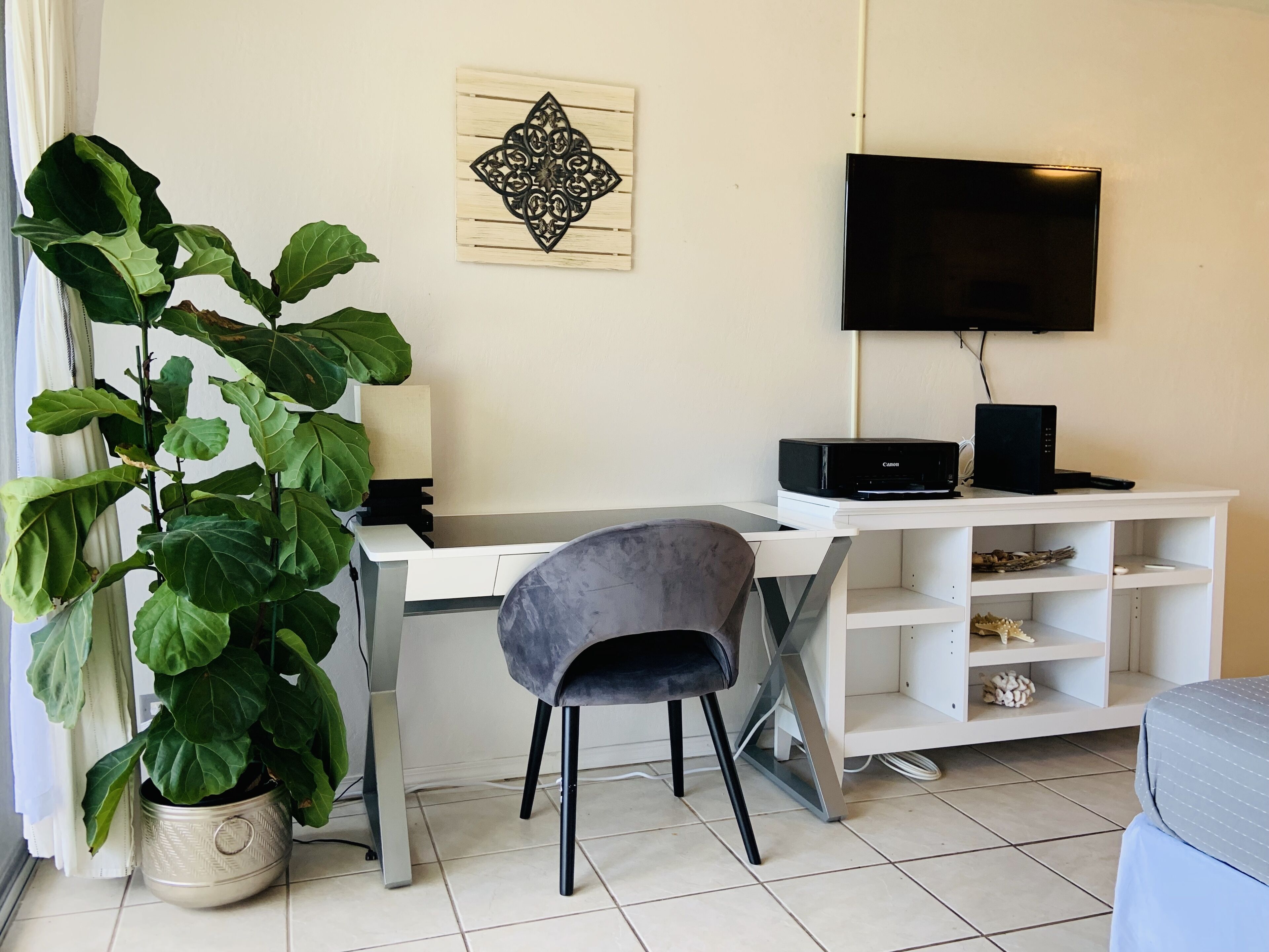 A modern living room with a white desk, a grey chair, a large potted plant, a flat-screen TV, and a white entertainment unit.