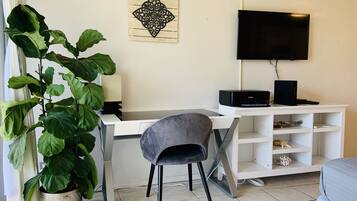 A modern living room with a white desk, a grey chair, a large potted plant, a flat-screen TV, and a white entertainment unit.
