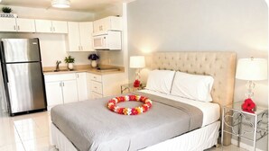 A hotel room with a bed, bedside lamps, a small table, a refrigerator, and a kitchen area. - RENOVATED Kona Studio By The Ocean!!! (Kailua)