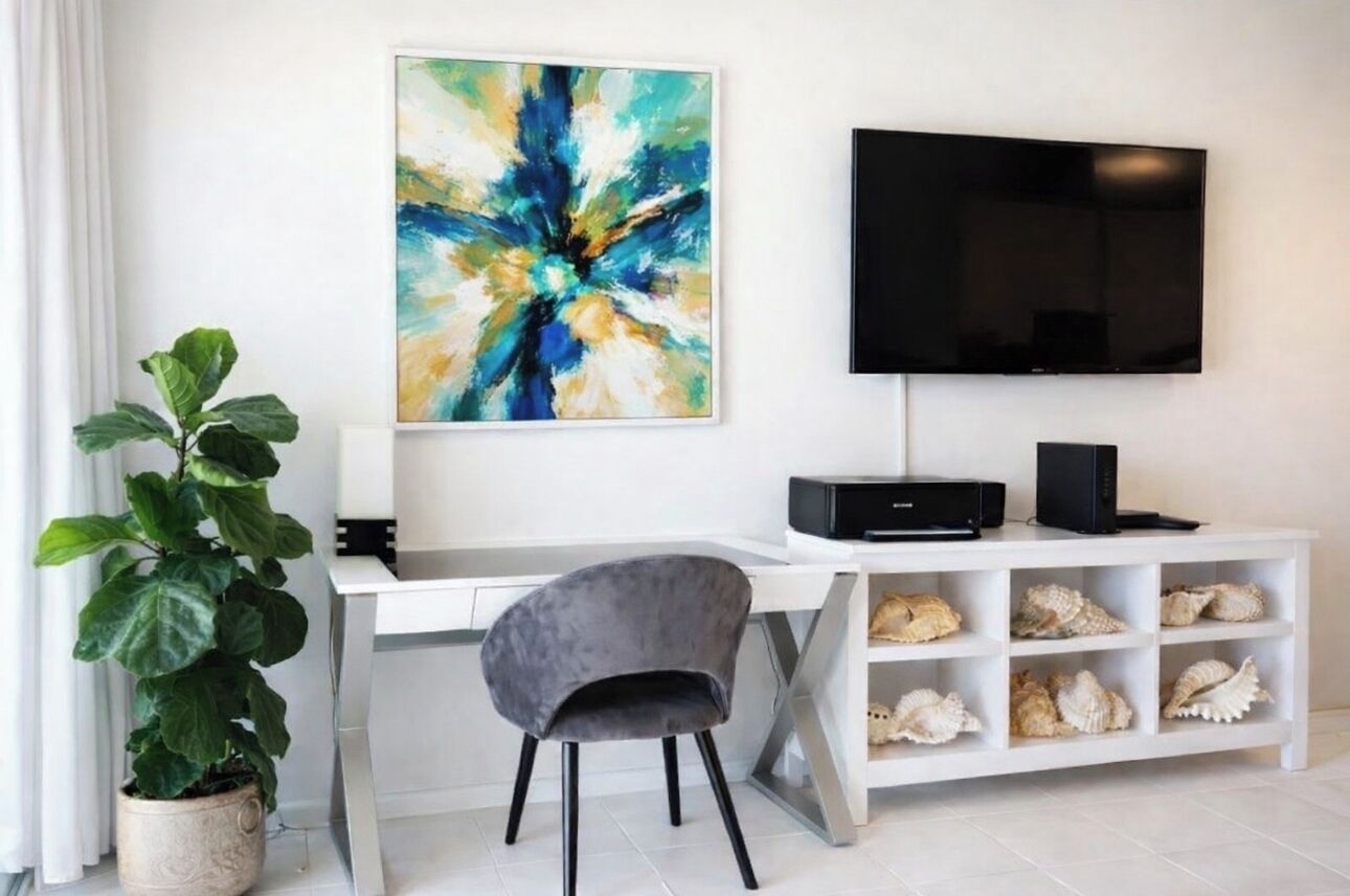 A modern room with a white desk, a grey chair, a large abstract painting, a flat-screen TV, and a shelf with seashells.