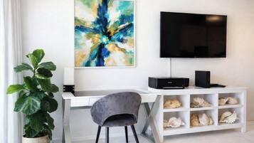 A modern room with a white desk, a grey chair, a large abstract painting, a flat-screen TV, and a shelf with seashells.