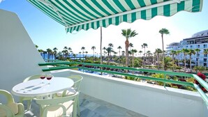 Un balcón con toldo a rayas verdes, una mesa redonda con dos copas de vino tinto y vistas a palmeras y un edificio al otro lado del agua.