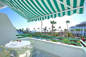Un balcone con un tendalino a righe verdi, un tavolo rotondo con due bicchieri di vino rosso e vista su palme e un edificio dall'altra parte dell'acqua.