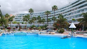 Una gran piscina con sillas de descanso y palmeras, rodeada por un edificio de hotel con varios balcones.