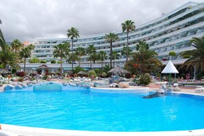 Una grande piscina con lettini e palme, circondata da un edificio alberghiero con numerosi balconi.