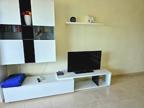 A modern living room with a flat-screen TV on a white entertainment unit, a remote control, and a blue towel on the floor.