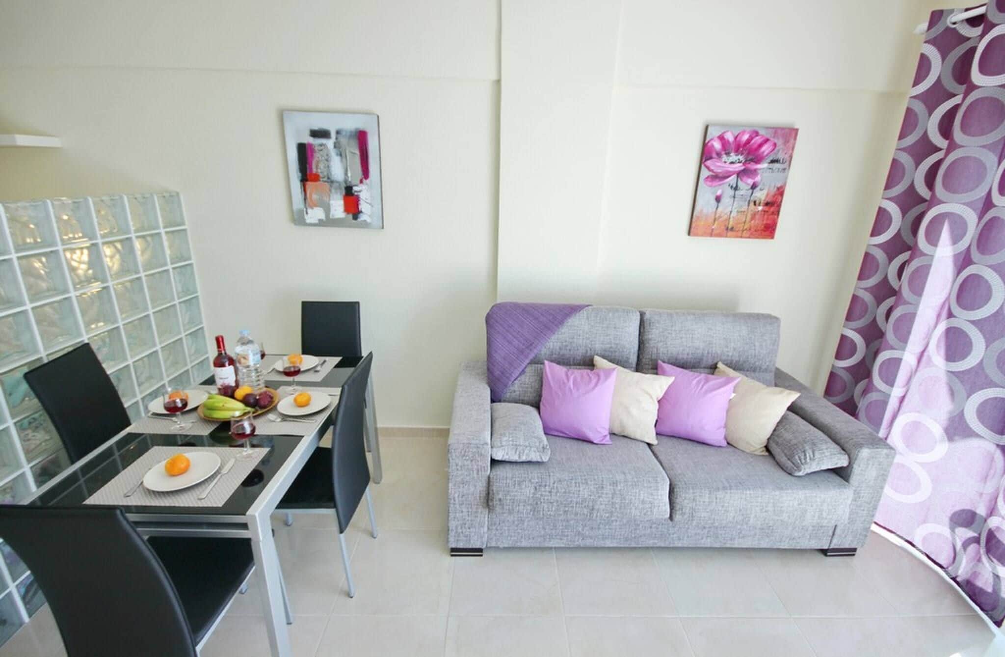 A modern living room with a grey sofa, a dining table with fruit and a bottle, and two wall art pieces.