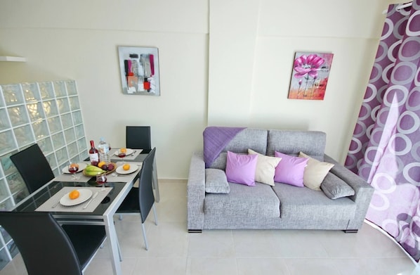 A modern living room with a grey sofa, a dining table with fruit and a bottle, and two wall art pieces.