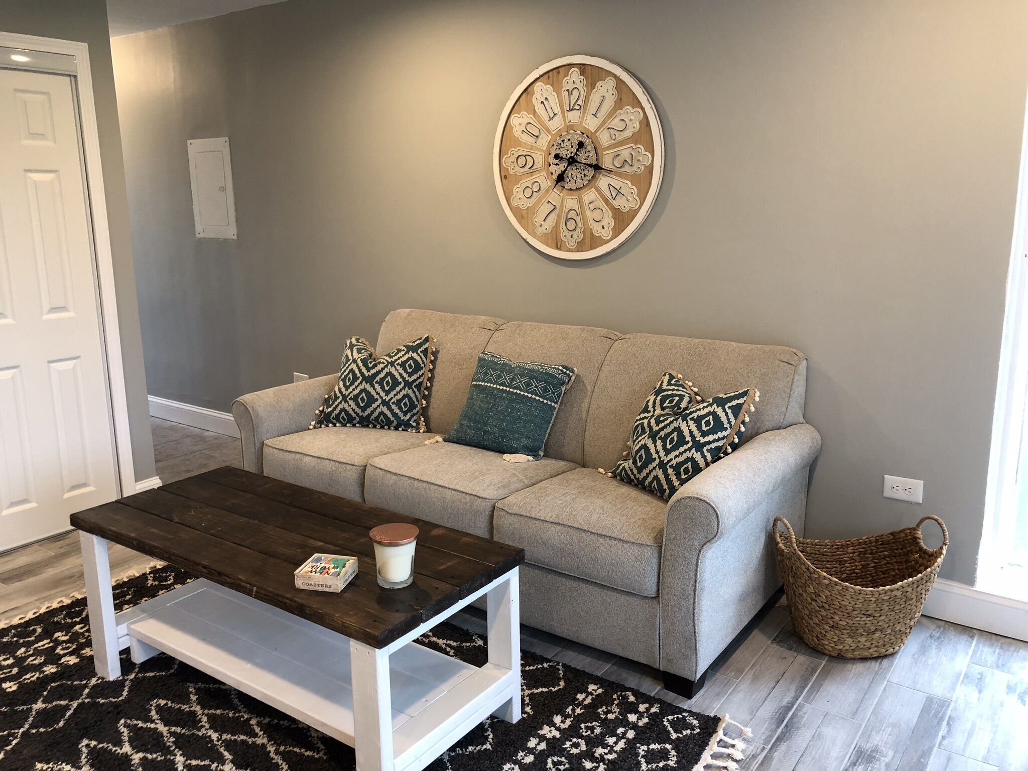 Ein Wohnzimmer mit grauem Sofa, gemusterten Kissen, einem Holztisch, einer Wanduhr und einem gewebten Korb.