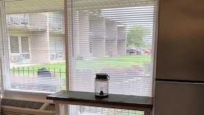 A kitchen with a refrigerator, two wooden stools, a window with blinds, and a view of a backyard.
