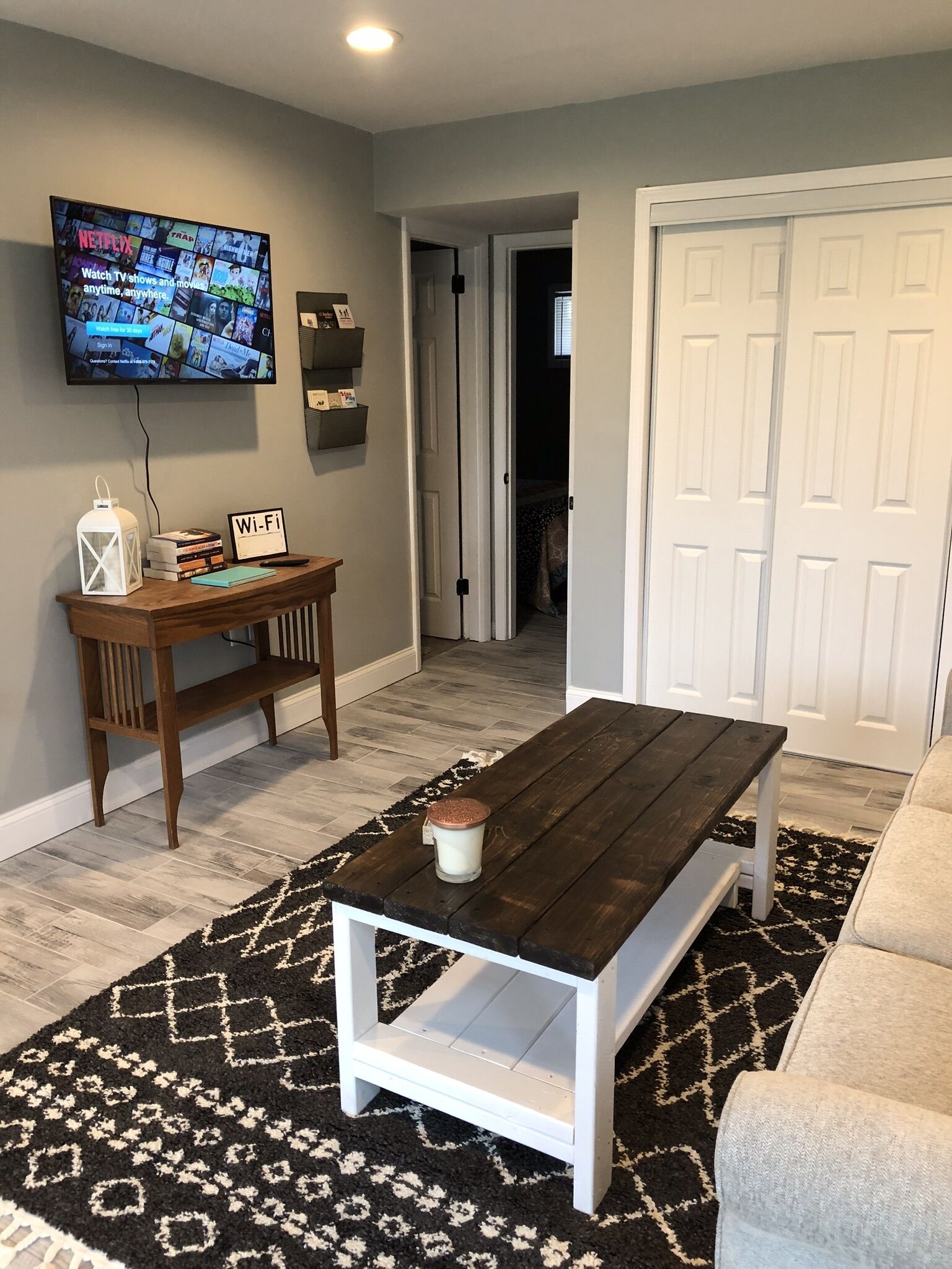 A living room with a dark wooden coffee table, a white bench, a TV mounted on the wall, and a door leading to another room.