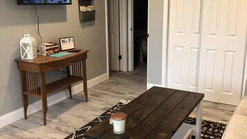 A living room with a dark wooden coffee table, a white bench, a TV mounted on the wall, and a door leading to another room.