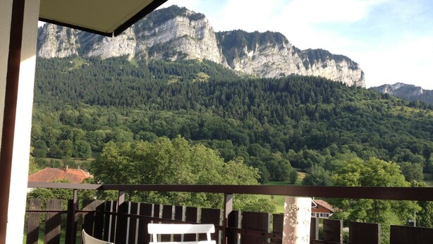Un balcon avec une table rouge et des chaises blanches, donnant sur un paysage montagneux parsemé de forêts denses.