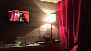 Une pièce avec un téléviseur diffusant une émission, un mur en bois, une lampe, une table sur laquelle se trouvent divers objets, et un rideau rouge.