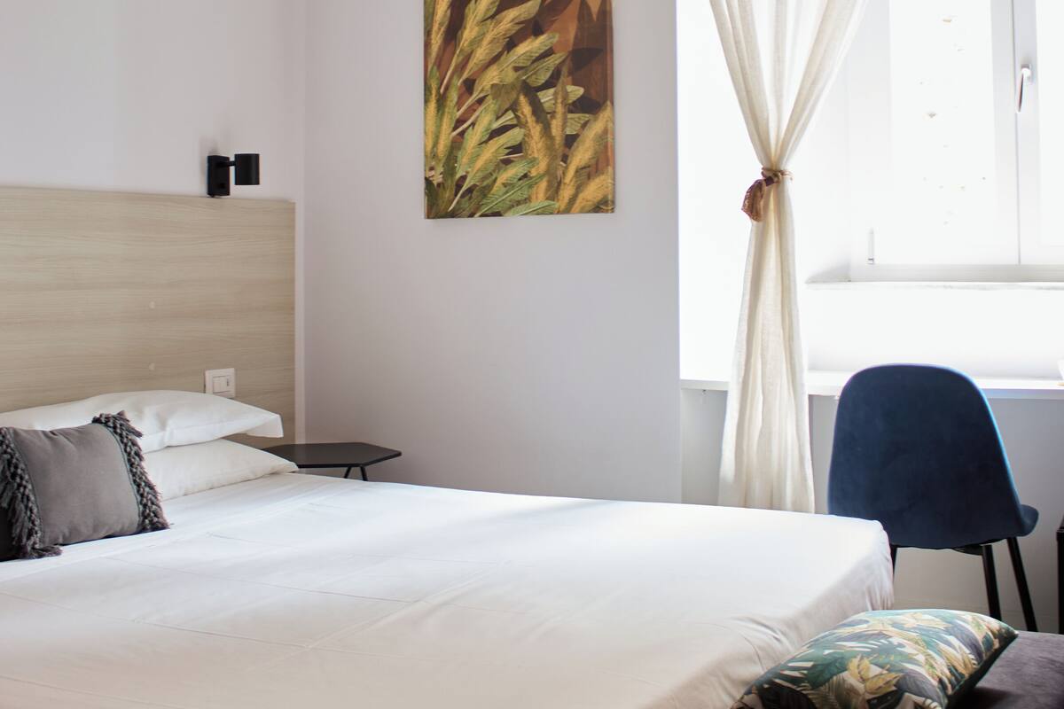 Ein Schlafzimmer mit Holzbalkendecke, einem Bett mit weißer Bettwäsche, einem blauen Sessel und einem Fenster mit weißen Vorhängen.