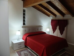 A bedroom with a red bedspread, two bedside lamps, a window with curtains, and a wooden ceiling beam. - B&B Eldorado (Sommacampagna)